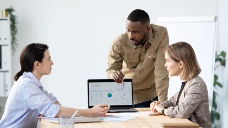 Three diverse professionals in business meeting examining analytics dashboard on laptop, standing around wooden conference table.