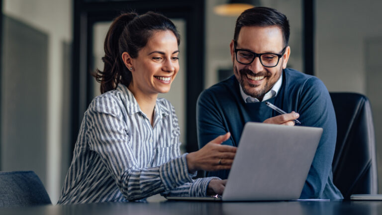 Two colleagues smiling while looking at a laptop together in an office setting, one holding a pen pointing at the screen.