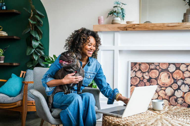 Woman with curly hair in denim outfit smiling, hugging dark French bulldog while using laptop on wicker table in cozy room with plants.
