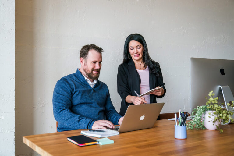 Two colleagues at wooden desk. Bearded man in blue sweater types on MacBook. Woman in black blazer takes notes. Workspace has monitor and plant.