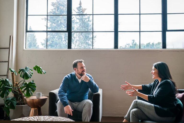 Two professionals in conversation: bearded man in blue sweater listening thoughtfully while woman in black blazer gestures expressively. Bright modern space with windows showing trees outside.
