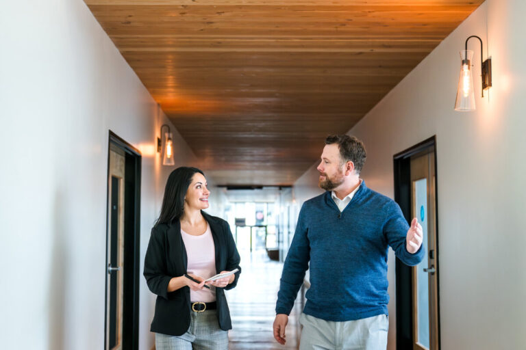 Two professionals conversing while walking: woman with notebook in black blazer and pink top smiles at bearded man in blue sweater gesturing. Modern hallway with wooden ceiling and natural light.