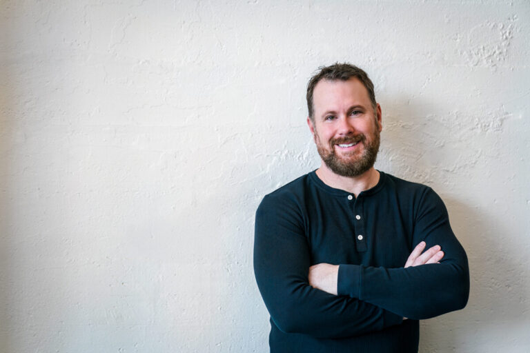 A bearded man with short brown hair smiles at camera while standing against white textured wall, wearing navy ribbed henley with arms crossed.