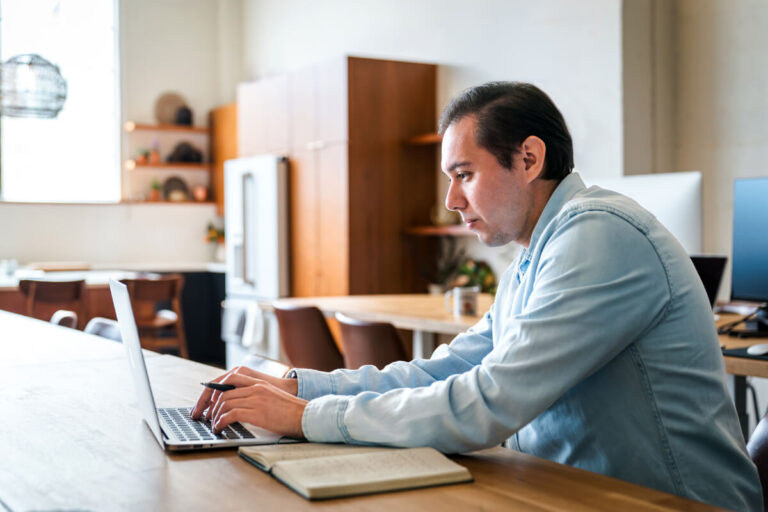 A dark-haired man in light blue shirt works intently on laptop at wooden table with notebook nearby. Modern workspace with wooden furniture and kitchen area visible.