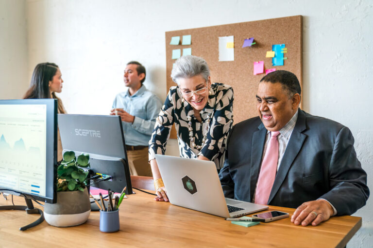 A man in dark suit and pink tie views laptop with gray-haired woman in patterned blouse while two colleagues converse behind them. Office with monitors and bulletin board.